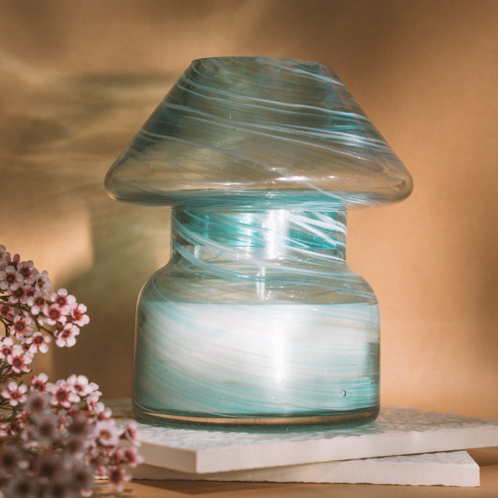 Mushroom candle lamp with aqua blue and gold glitter swirls on clear glass. Mushroom lamp is filled with 100% soy wax placed on white tiles and tan background.