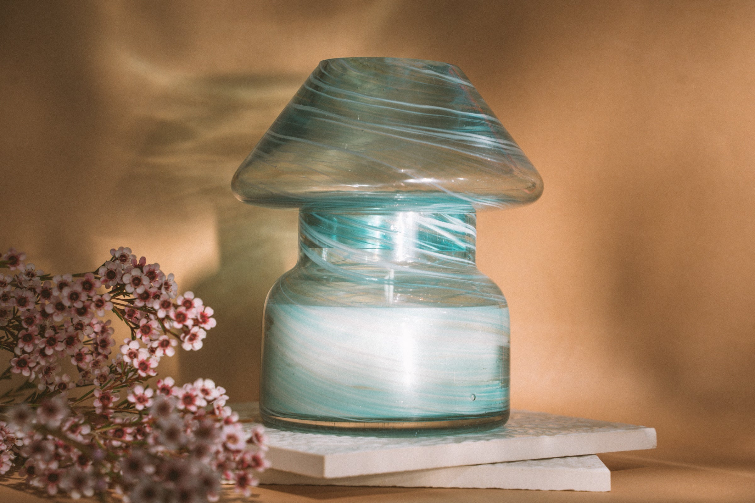Mushroom candle lamp with aqua blue and gold glitter swirls on clear glass. Mushroom lamp is filled with 100% soy wax placed on white tiles and tan background.