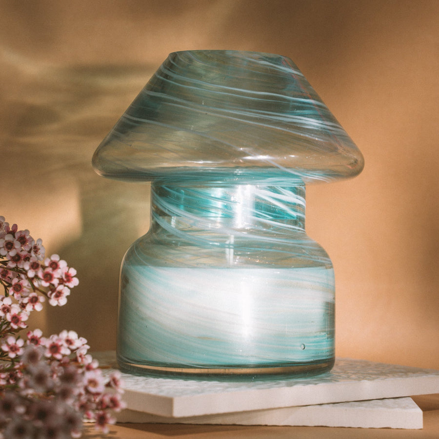 Mushroom candle lamp with aqua blue and gold glitter swirls on clear glass. Mushroom lamp is filled with 100% soy wax placed on white tiles and tan background.