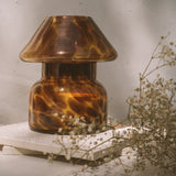 Mushroom candle lamp with light and dark brown spots on tan coloured glass. Leopard candle lamp is filled with 100% soy wax placed on white tiles with dried flowers.