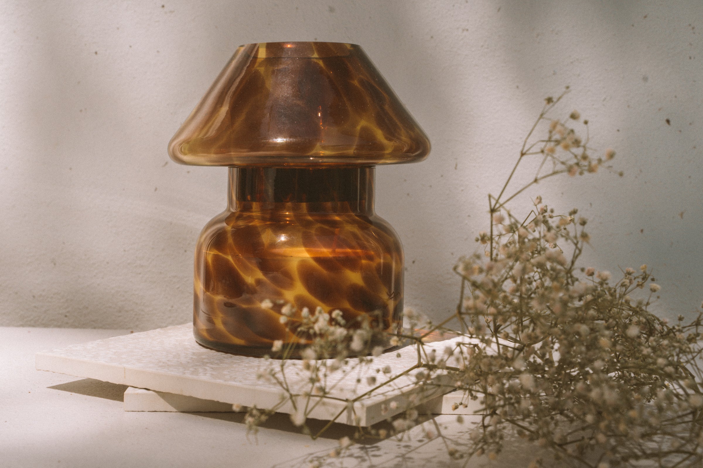 Mushroom candle lamp with light and dark brown spots on tan coloured glass. Leopard candle lamp is filled with 100% soy wax placed on white tiles with dried flowers.