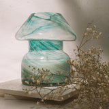 Mushroom candle lamp with aqua blue and gold glitter swirls on clear glass. Mushroom lamp is filled with 100% soy wax sitting on white tiles with dried flowers.