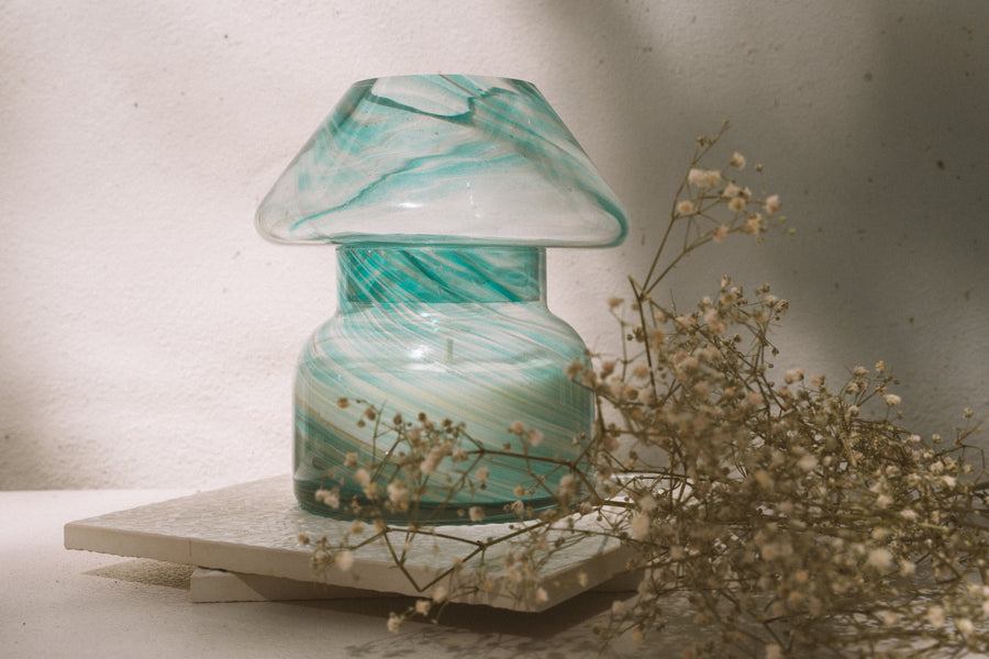 Mushroom candle lamp with aqua blue and gold glitter swirls on clear glass. Mushroom lamp is filled with 100% soy wax sitting on white tiles with dried flowers.