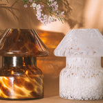 Mushroom candle lamp with light and dark brown spots on tan coloured glass. Leopard candle lamp is lit next to white candle lamp on tan background with dried flowers.
