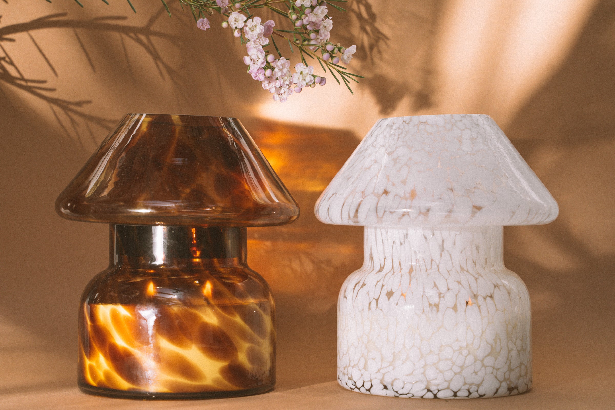 Mushroom candle lamp with light and dark brown spots on tan coloured glass. Leopard candle lamp is lit next to white candle lamp on tan background with dried flowers.
