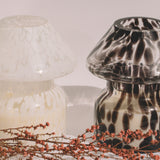 Mushroom candle lamp with black spots on clear glass next to white candle lamp. Filled with 100% soy wax with dried flowers.