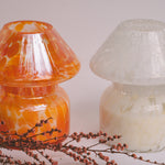 Mushroom candle lamp with light and dark orange spots on clear glass with opal tint next to white candle lamp. Filled with 100% soy wax displayed with dried flowers.