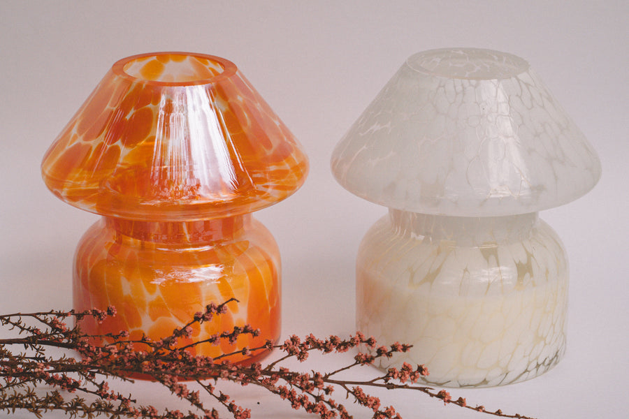 Mushroom candle lamp with light and dark orange spots on clear glass with opal tint next to white candle lamp. Filled with 100% soy wax displayed with dried flowers.