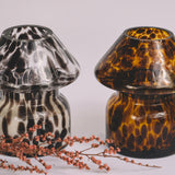 Mushroom candle lamp with black spots on clear glass next to leopard print candle lamp. Filled with 100% soy wax with dried flowers.