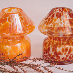 Mushroom candle lamp with light and dark orange spots on clear glass with opal tint next to retro coloured candle lamp. Filled with 100% soy wax.