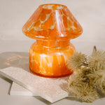 Mushroom candle lamp with light and dark orange spots on clear glass with opal tint. Filled with 100% soy wax on tiles with dried flowers.