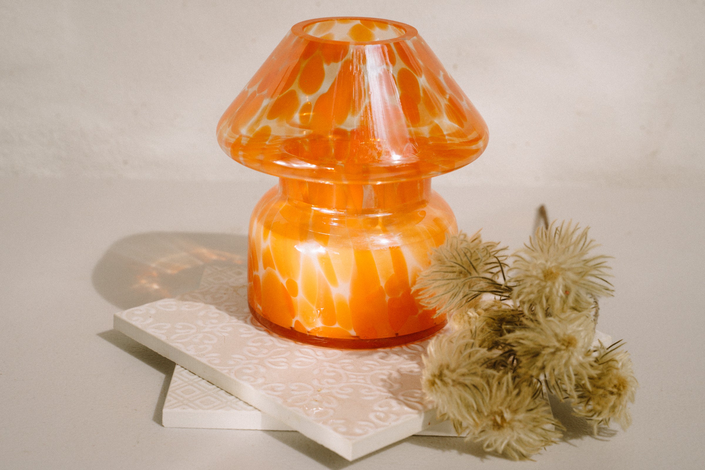 Mushroom candle lamp with light and dark orange spots on clear glass with opal tint. Filled with 100% soy wax on tiles with dried flowers.