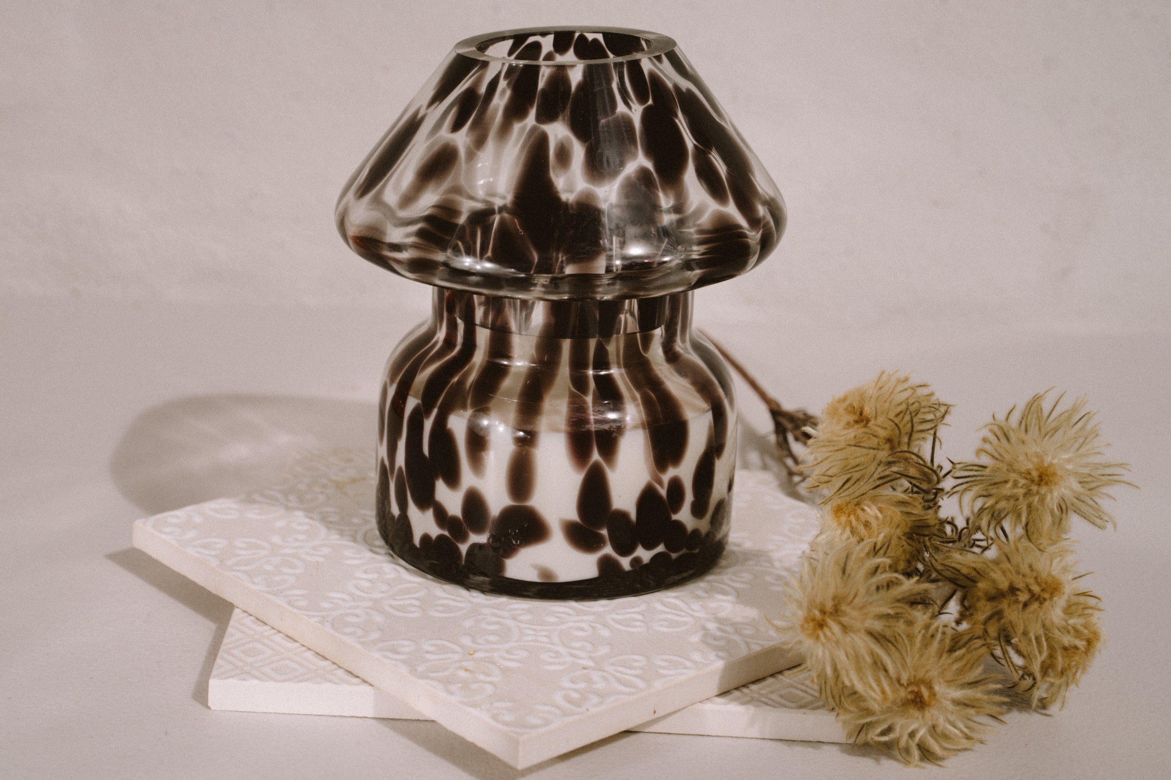 Mushroom candle lamp with black spots on clear glass. Filled with 100% soy wax on tile with dried flowers.