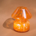 Mushroom candle lamp with light and dark orange spots on clear glass with opal tint. Filled with 100% soy wax, candle lamp is on tan background.