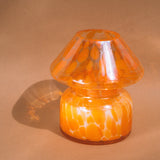 Mushroom candle lamp with light and dark orange spots on clear glass with opal tint. Filled with 100% soy wax, candle lamp is on tan background.