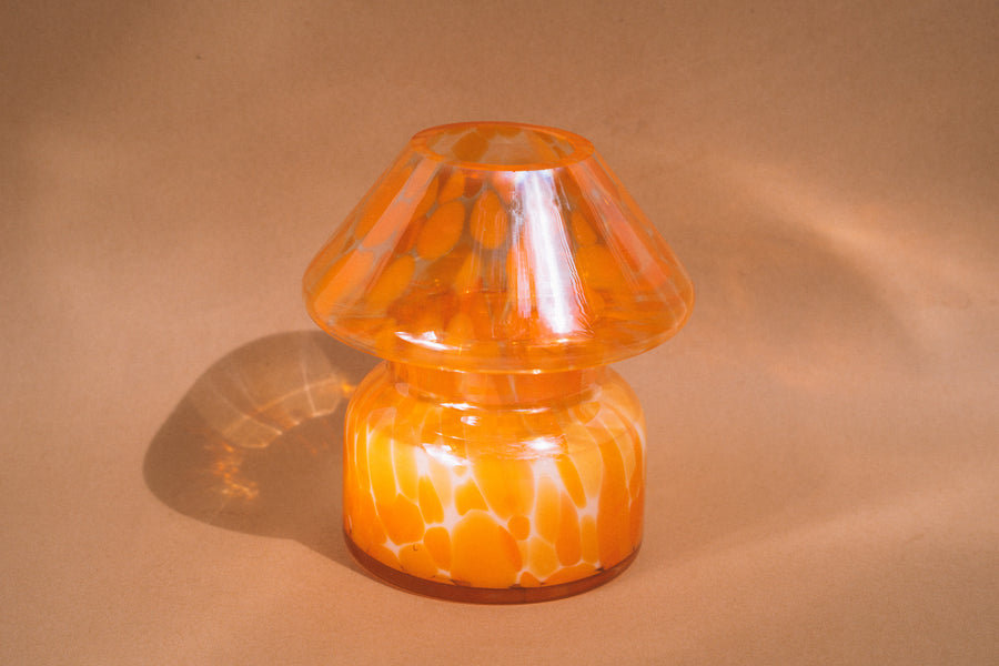 Mushroom candle lamp with light and dark orange spots on clear glass with opal tint. Filled with 100% soy wax, candle lamp is on tan background.