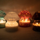 Mushroom candle lamp with white spots on clear glass. Candle lamp is lit next to 3 different coloured mushroom lamps.