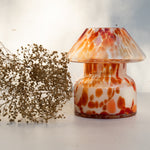 Mushroom candle lamp with red, orange and white spots on clear glass. Retro lamp is filled with 100% soy wax placed next to dried flowers.