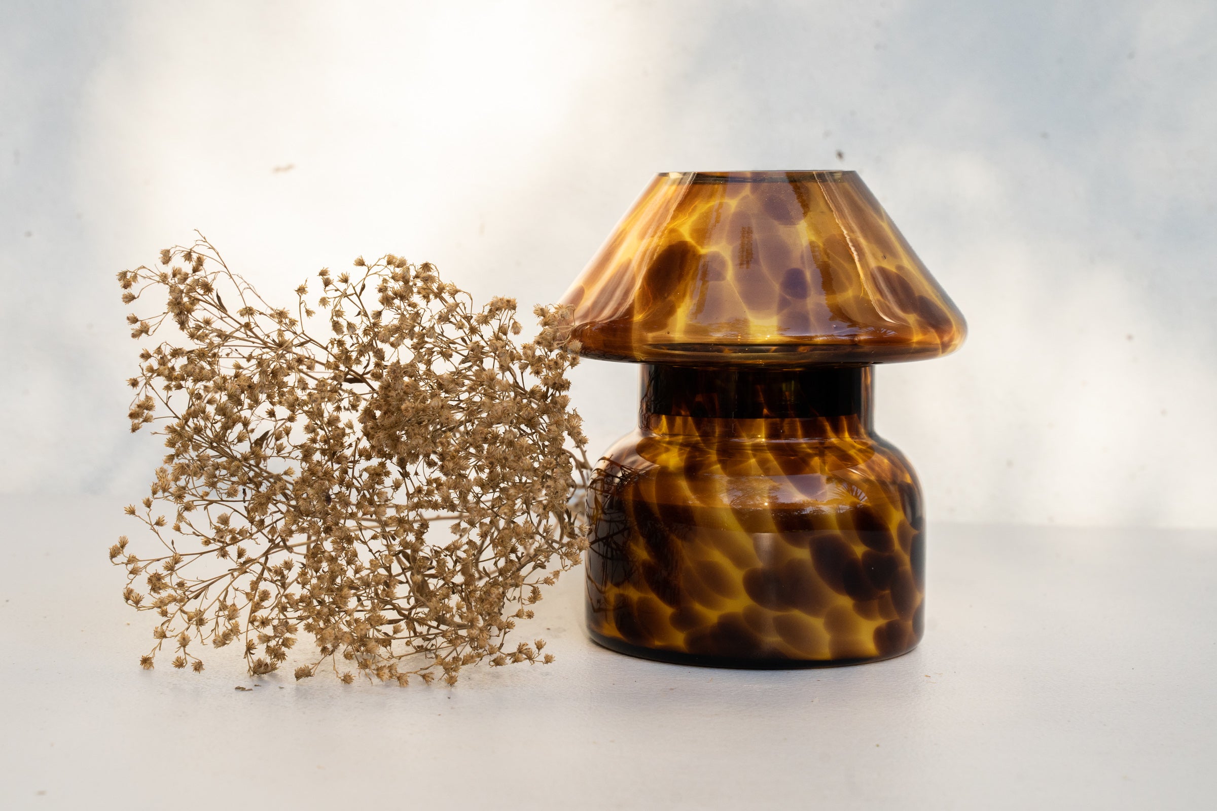 Mushroom candle lamp with light and dark brown spots on tan coloured glass. Leopard candle lamp is filled with 100% soy wax next to dried flowers.